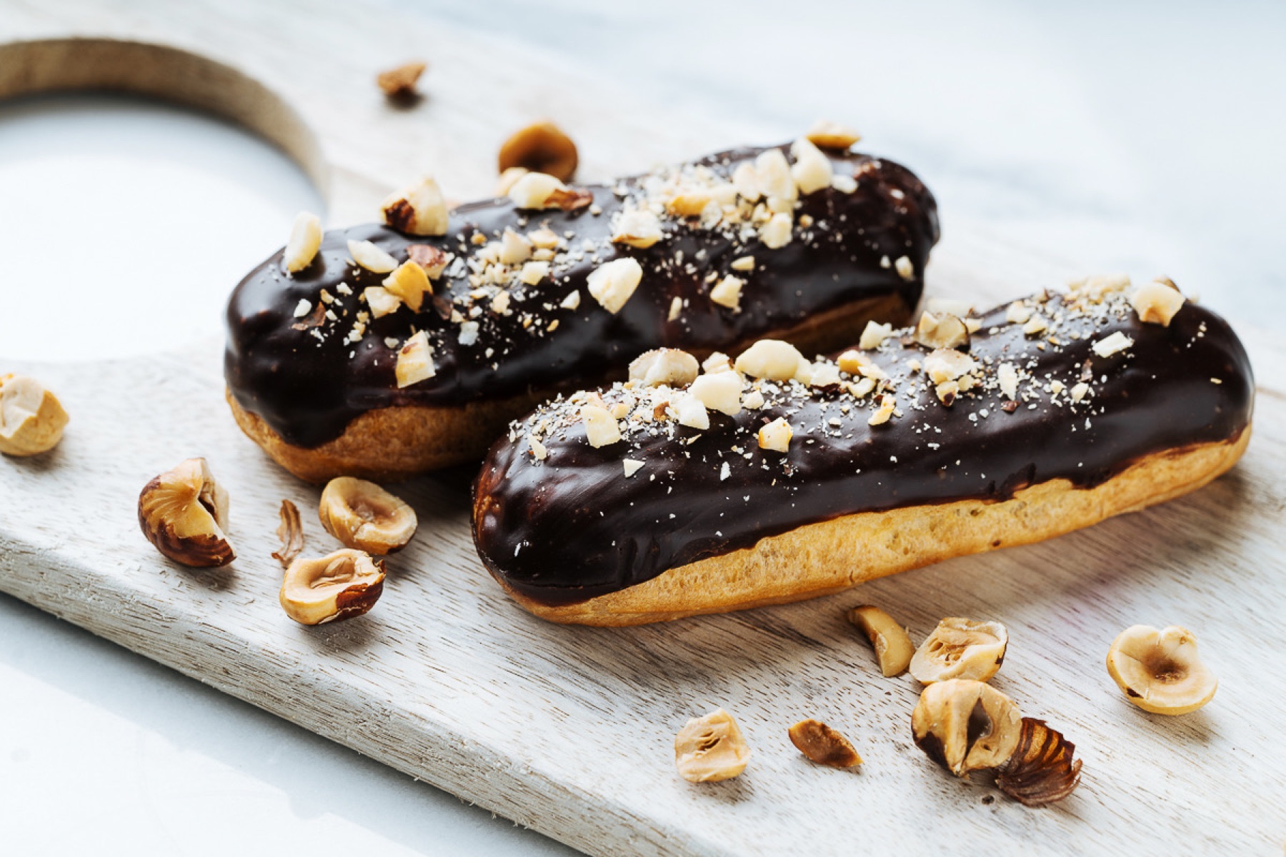 Gluten-free chocolate eclairs topped with crushed hazelnuts on a pale wooden board.